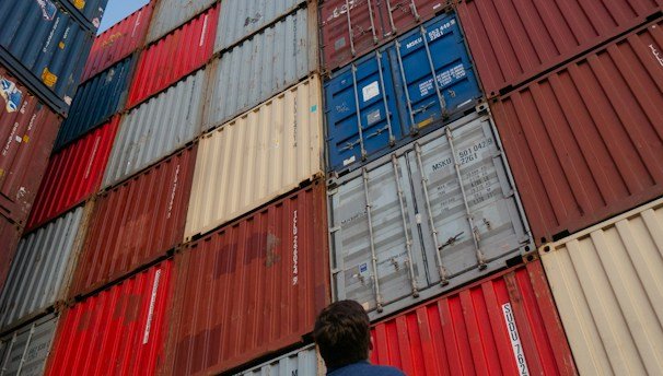man in black jacket standing in front of red and blue intermodal containers