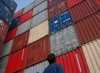 man in black jacket standing in front of red and blue intermodal containers